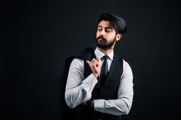 Portrait of a young handsome successful Indian in a classic black three-piece business suit on a dark background, Holding his jacket over his shoulder