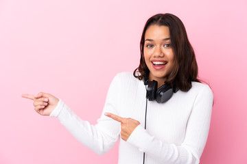 Young woman with earphones over isolated pink wall surprised and pointing side