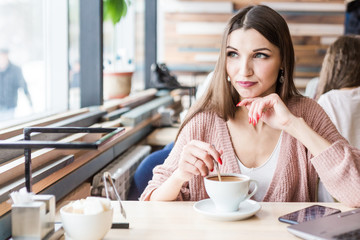 Beautiful young woman sits at a table in a cafe with a cup of coffee and looks out the window
