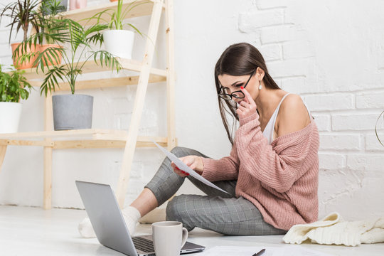 A young happy woman sits on the floor in a bright apartment or office interior and works at a laptop, freelancer girl at work, the concept of distance learning students, paperwork and online learning