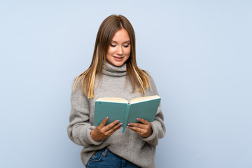 Fototapeta premium Teenager girl with sweater over isolated blue background holding and reading a book
