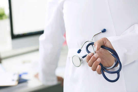 Rear view image of doctor with arms behind back holding stethoscope and looking at pc screen. Selective focus on stethoscope. Health and medical concept.