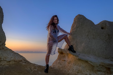 Naklejka premium Silhouette of a young and sexy girl. She stands on a mountain and poses, dressed in a cloak and black boots against the sunset. Happy celebration victory success woman
