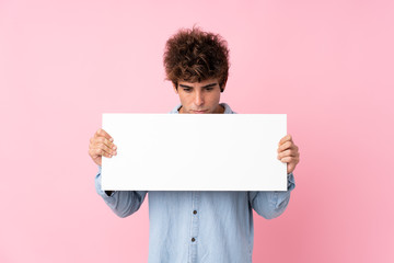 Young caucasian man over isolated pink background holding an empty white placard for insert a...