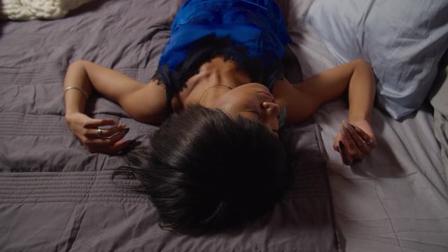 Exhausted overloaded lovely multiethnic female in blue pajamas falling down on cozy bed late at night, being jaded and fatigued. Tired pretty young woman in nightwear falling asleep in her bedroom.
