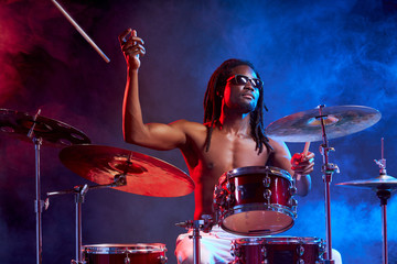 portrait of talented african man playing on drums, with naked skin wearing eyeglasses isolated over smoky neon background