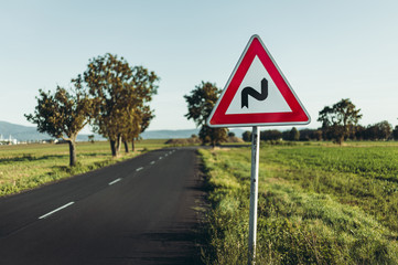 Photo of illuminated and warning road sign - double curve next to the road on meadow. Double Bend warning road sign on country road with trees and beautiful landscape on background - European sign.