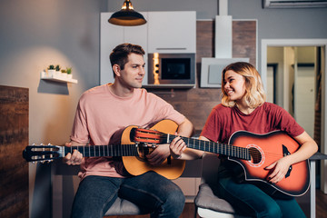 Nice couple of lovely people play the guitar while sitting in bedroom, wearing casual clothes, performing music at home