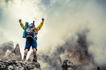 Young man hiker standing on stone mountain with raised up arms with cloudy sky and fog. Yellow...