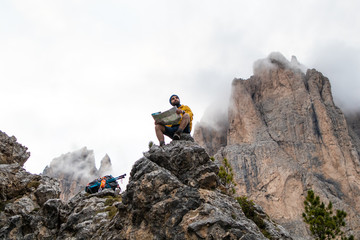 Young man hiker sitting on stone mountain reading map, with cloudy sky and fog. Yellow jacket,...