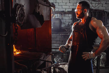 Fototapeta na wymiar Portrait of a professional brutal caucasian blacksmith with strong muscles, side view on man opposite of fire