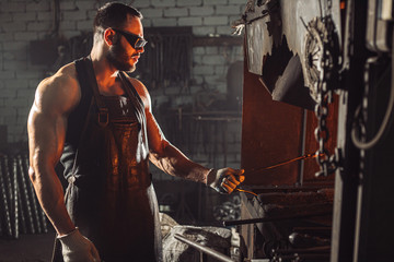 Fototapeta na wymiar bearded man in good physical form with strong muscles warming iron for manufacturing it, wearing leather apron and protective eyeglasses isolated in workshop