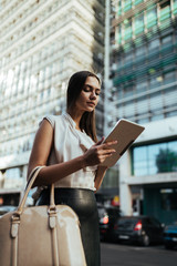 slender tanned girl in a light blouse looks at a tablet pc against the background of tall buildings