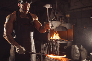 muscular adult with beard wearing leather apron working in workshop, heating. hardened metal in...