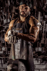 Fototapeta na wymiar portrait of young muscular strong caucasian man blacksmith looking at camera, wearing leather apron uniform, holding hammer isolated in dark space, workshop