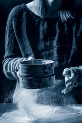 Process of making homemade bread dough. Woman hands sifts flour from vintage sieve on dark kitchen. Black table with flour. Home bread baking. Photo series. Color of the year 2020 classic blue toned