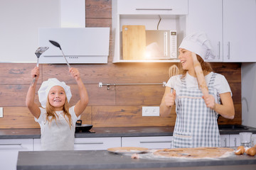 caucasian mother and daughter dance while baking in kitchen. holding kitchen utensils, kitchenware