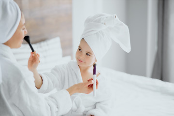 young caucasian woman sit in towel and bathrobe with daughter hold makeup brushes, white bedroom