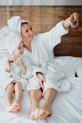 Obraz na płótnie Canvas lovely woman and her daughter wearing bathrobe and towel lie on bed and take photo of themselves, white bedroom, friendly family use smartphone