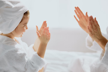 Fototapeta premium family game at home. friendly beautiful mother and her daughter sit clapping hands, play with each other, wearing white bathrobe and towel