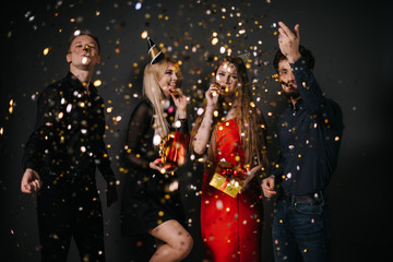 Obraz na płótnie Canvas Two cheerful guys blow a festive whistle and throw up a colorful confetti. Two beautiful happy girl are holding bright gift boxes in hands. Shooting in professional studio on isolated black background