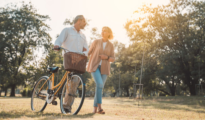 Beautiful Senior couple walking their bike along happily talking in park.mature couple in summer park.Elderly man and old woman with bicycles outside in spring nature.