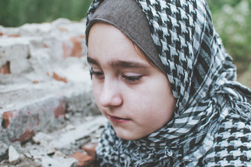 a refugee child in the war, a Muslim girl with a dirty face on the ruins, the concept of peace and war, the child is crying and waiting for help. - obrazy, fototapety, plakaty