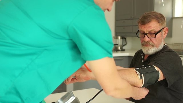 Young doctor or nurse measures the blood pressure of an elderly man in his home, in the kitchen. Emergency ambulance