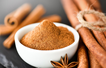 Cinnamon sticks spices on dark stone table. Cinnamons milled powder in white bowl on black board.