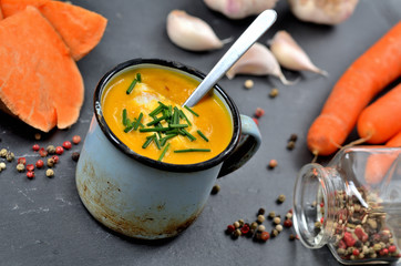 Sweet potato soup with cream and fresh chive in old enamel cup. Garlic, pumpkin and half pieced carrot in background. Slate plate and spoon.