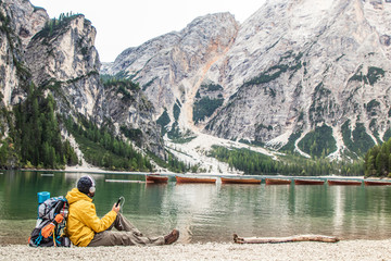Young tourist hiker man listening to music sitting by alpine lake Lago Di Braies (Pragser wildsee) in Trentino, Dolomites mountains, Italy.