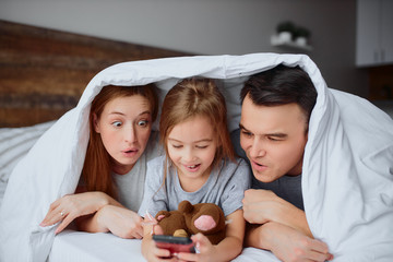 happy funny parents and daughter lying on bed and watching video together on smartphone, under white blanket