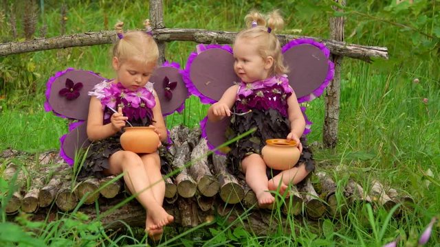 Adorable girls with butterfly wings eat honey from pots with a spoon. Children pretend to be purple butterflies
