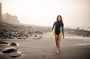 Fit girl in the black diving suit walking on the shore holding a yellow surfing board in hands