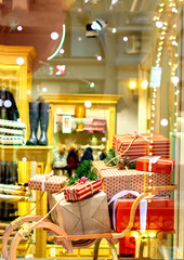 Boxes of Christmas gifts wrapped in kraft paper with ribbons and red bows on a retro wooden sleigh lie on top of each other in a clothing store window with bokeh lights. New Year background.