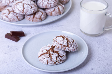 Chocolate cookies with cracks, strewed with icing sugar. Homemade baking. Background: a cup of milk, chocolate and a plate of cookies. Selective focus, close-up.