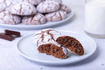 Chocolate cookies with cracks, strewed with icing sugar. Homemade baking. Background: a cup of milk, chocolate and a plate of cookies. Selective focus, close-up.