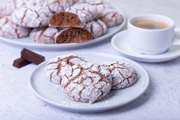 Chocolate cookies with cracks, strewed with icing sugar. Homemade baking. Background: a cup of coffee, chocolate and a plate of cookies. Selective focus, close-up.