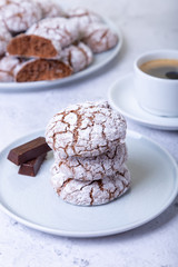 Chocolate cookies with cracks, strewed with icing sugar. Homemade baking. Three cookies in a stack. Background: a cup of coffee and a plate with cookies. Selective focus, close-up.