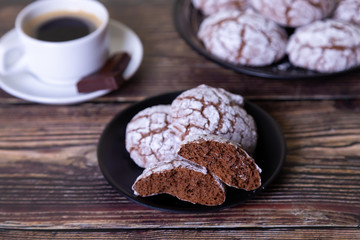 Chocolate cookies with cracks, strewed with icing sugar. Homemade baking, wooden background. Background: a cup of coffee, chocolate and a plate of cookies. Selective focus, close-up.