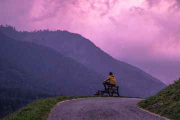 Young man sitting on a bench in front of the mountains after sunset with purple sky. Dolomites,South Tyrol, Italy
