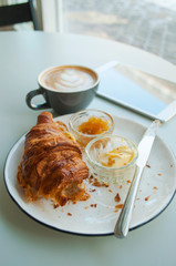 Fresh croissant with jam and butter and cappuccino with beautiful latte art on grey table in a cafe.