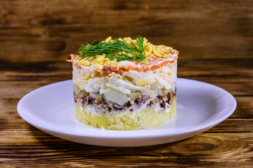 Plate with traditional russian salad Mimosa on a wooden table. Layered salad with potatoes, sardine, cheese, carrot, eggs and mayonnaise