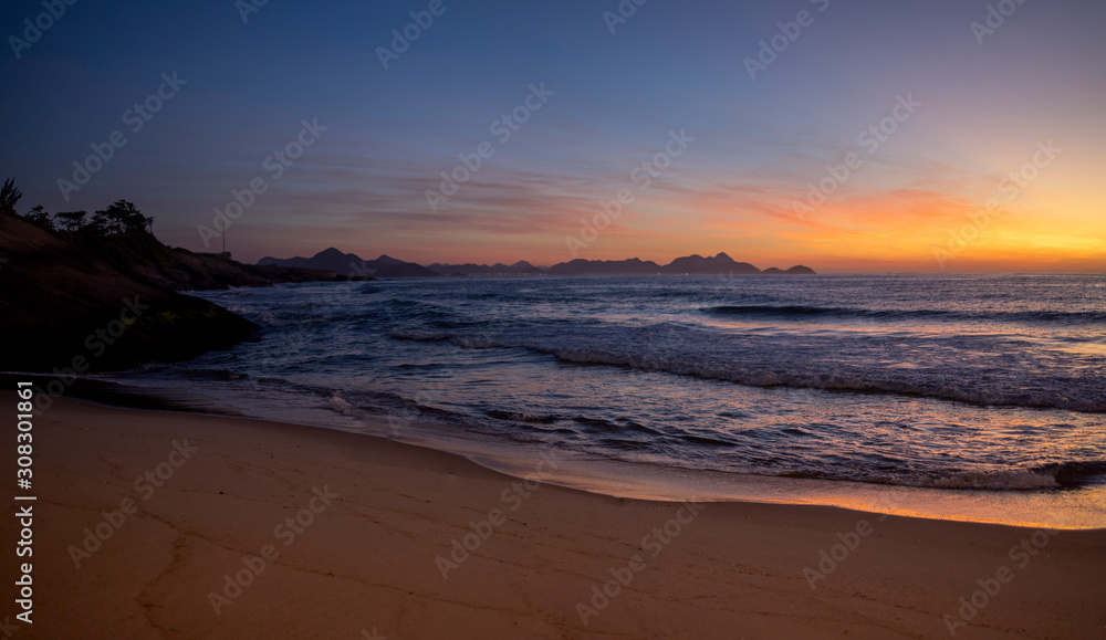 Wall mural Warm colourful glowing sunrise at the crack of dawn at the Arpoador Devil's beach in Rio de Janeiro with waves coming in reflecting the orange colours of the sun and a pristine sand beach