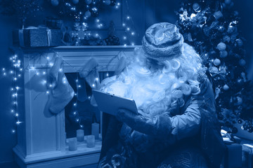 Russian Santa Claus (Grandfather Frost)  use his mobile tablet with glowing screen to choose  gifts for Christmas and New Year. Classic blue color 2020