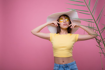 Asian girl in a hat and in yellow glasses stands next to a home palm holds a hat in a yellow T-shirt and on a pink background while waiting for travel