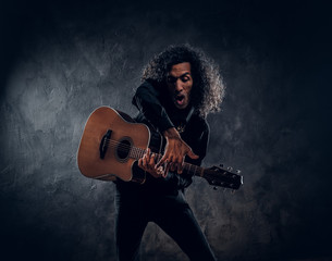 Middle aged emotional singer is playing an acoustic guitar at dark studio.