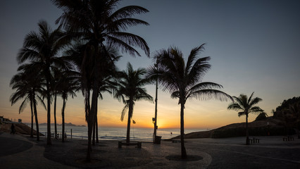 Fototapeta na wymiar Panorama of a sunrise on the Arpoador boulevard with the Devils beach and silhouetted palm trees in Rio de Janeiro against a clear orange and blue sky