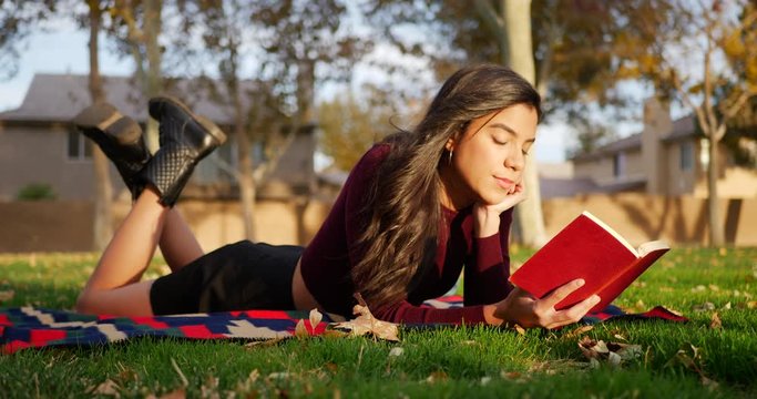 Attractive young hispanic woman college student reading a book outdoors in the park before class starts in the fall semester.