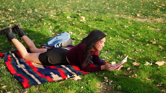 A beautiful adult woman student reading a book and relaxing in the park before her college literature class in autumn.